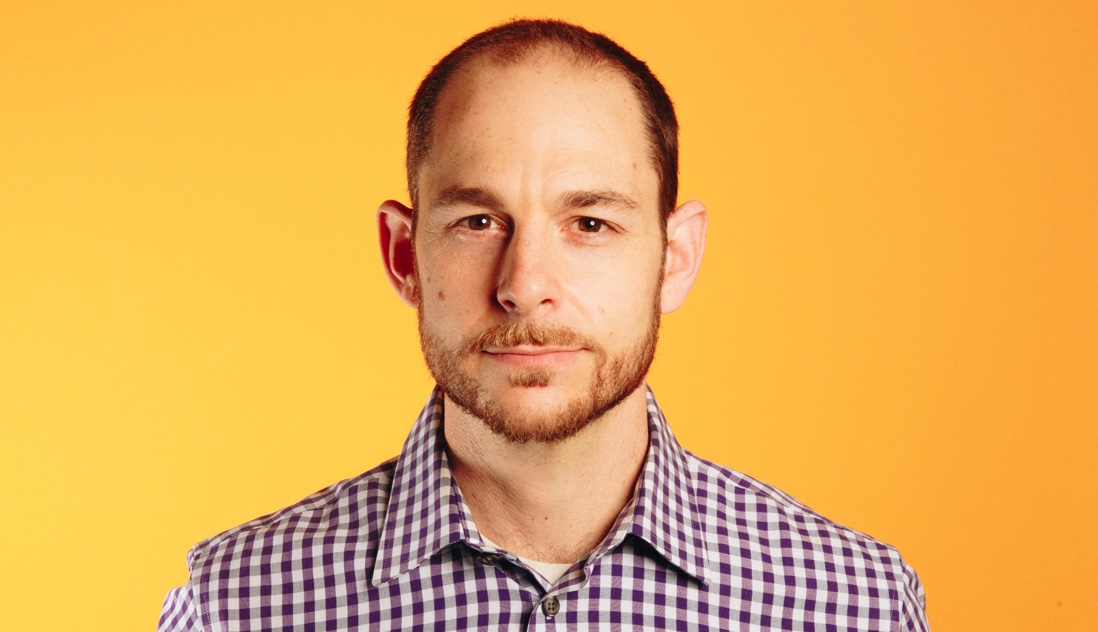 Portrait of Brian Rosenbaum, a leader who uses NationBuilder, against a yellow background, wearing a purple and white checked shirt.