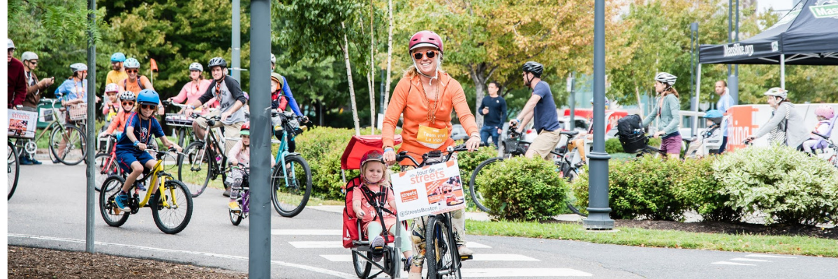 three people riding bicycles along a tree-lined path. the woman at the front of the line is wearing an orange sweater and has a child seat on her bicycle with a small child in it