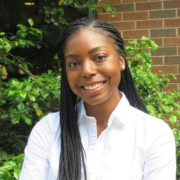 Ambar, a young black woman with her hair braided in long, thin cornrows, smiles at the camera while standing in front of green foliage and a brick wall.