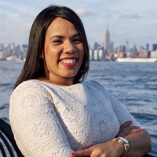 Anabelle, a Latina woman with shoulder-length straightened brown hair wearing a white shirt, smiles at the camera while standing in front of the boston harbor with the city's skyline in the distance.