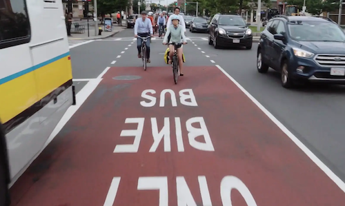 Allston Bus-Bike Lane video A photograph of the bus-bike lane, with three cyclists riding along it towards the viewer, with cars on their left and a bus on their right.