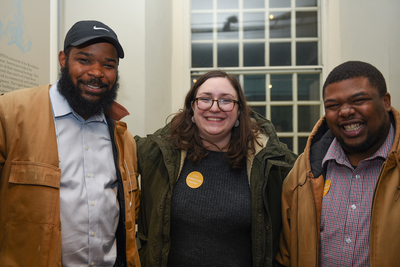 Three people, two Black men and a White woman, stand arm in arm and give big smiles to the camera.