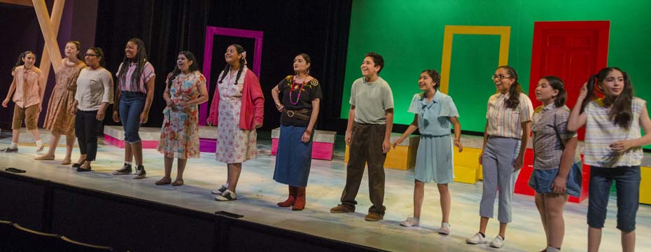A line of youth actors stand across the front of the stage during a scene from The House on Mango Street