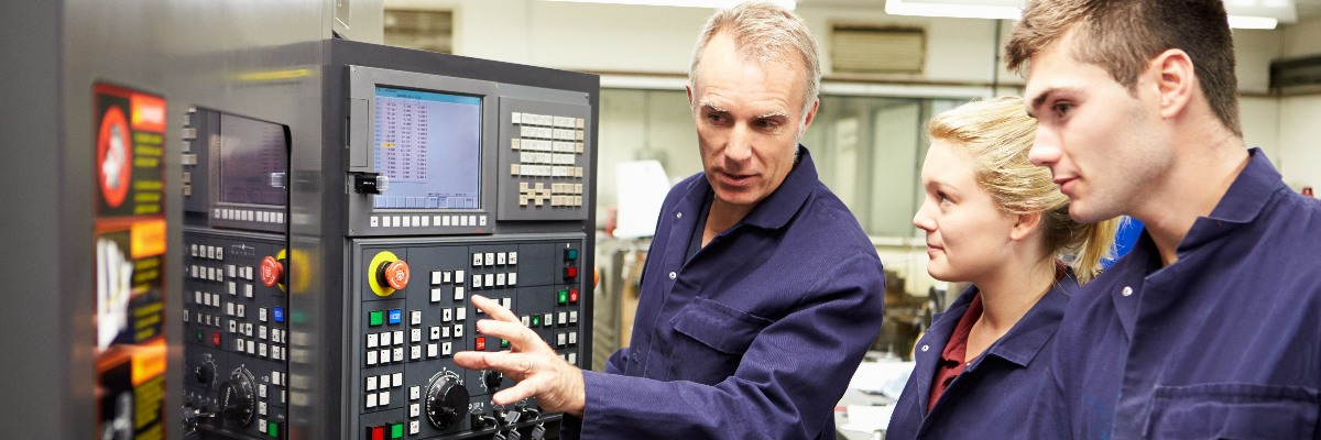 Two apprentices looking at a machine, at which their instructor is pointing.