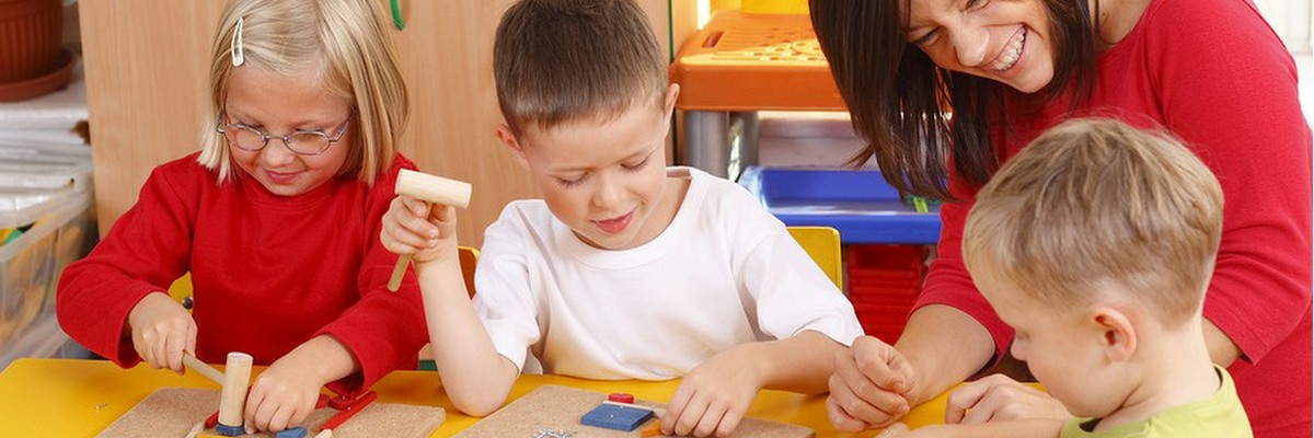 Three young children playing at a table, supervised by a smiling adult.