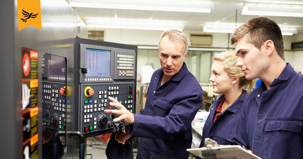 Two apprentices looking at a machine, at which their instructor is pointing.