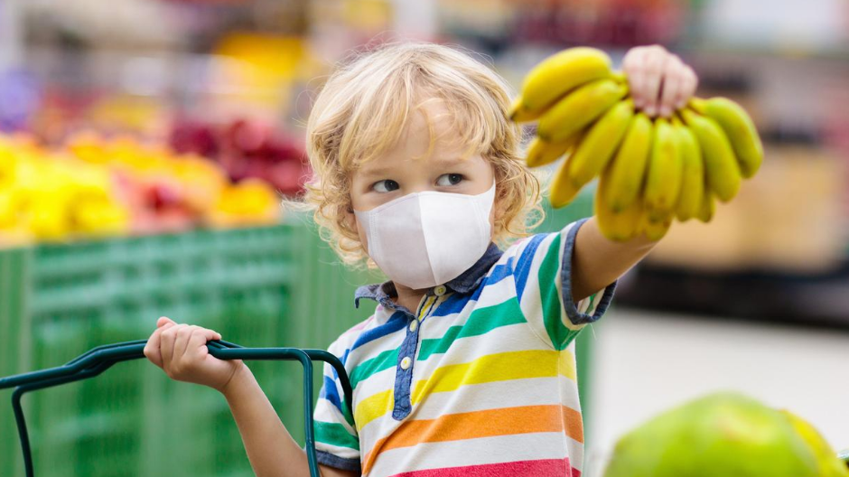 A picture of a little boy in grocery store wearing a mask and holding a bunch of bananas.